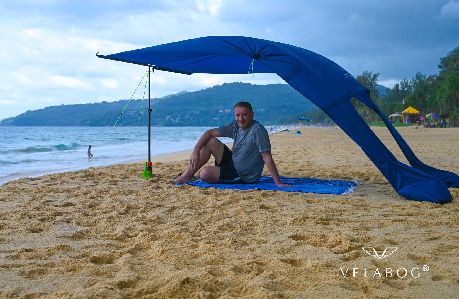 Velabog «Breeze» żagiel słoneczny - ciemnoniebieski. Najlepsza ochrona przeciwsłoneczna na plaży w Tajlandii nawet podczas deszczu. Może być ustawiony przy lekkim lub silnym wietrze. Widok z boku. Osoby.