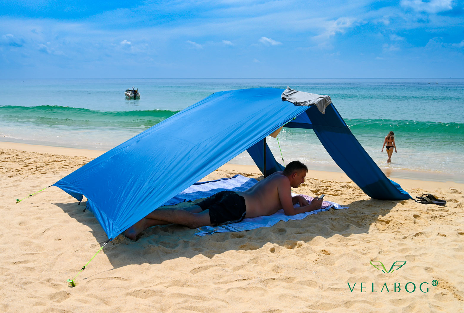 Velabog «Breeze» żagiel słoneczny - niebieski. Przewiewna ochrona przeciwsłoneczna na plażę nawet bez wiatru. Opcja używania: bez wiatru lekki lub porywisty wiatr. Widok z boku. Osoby.