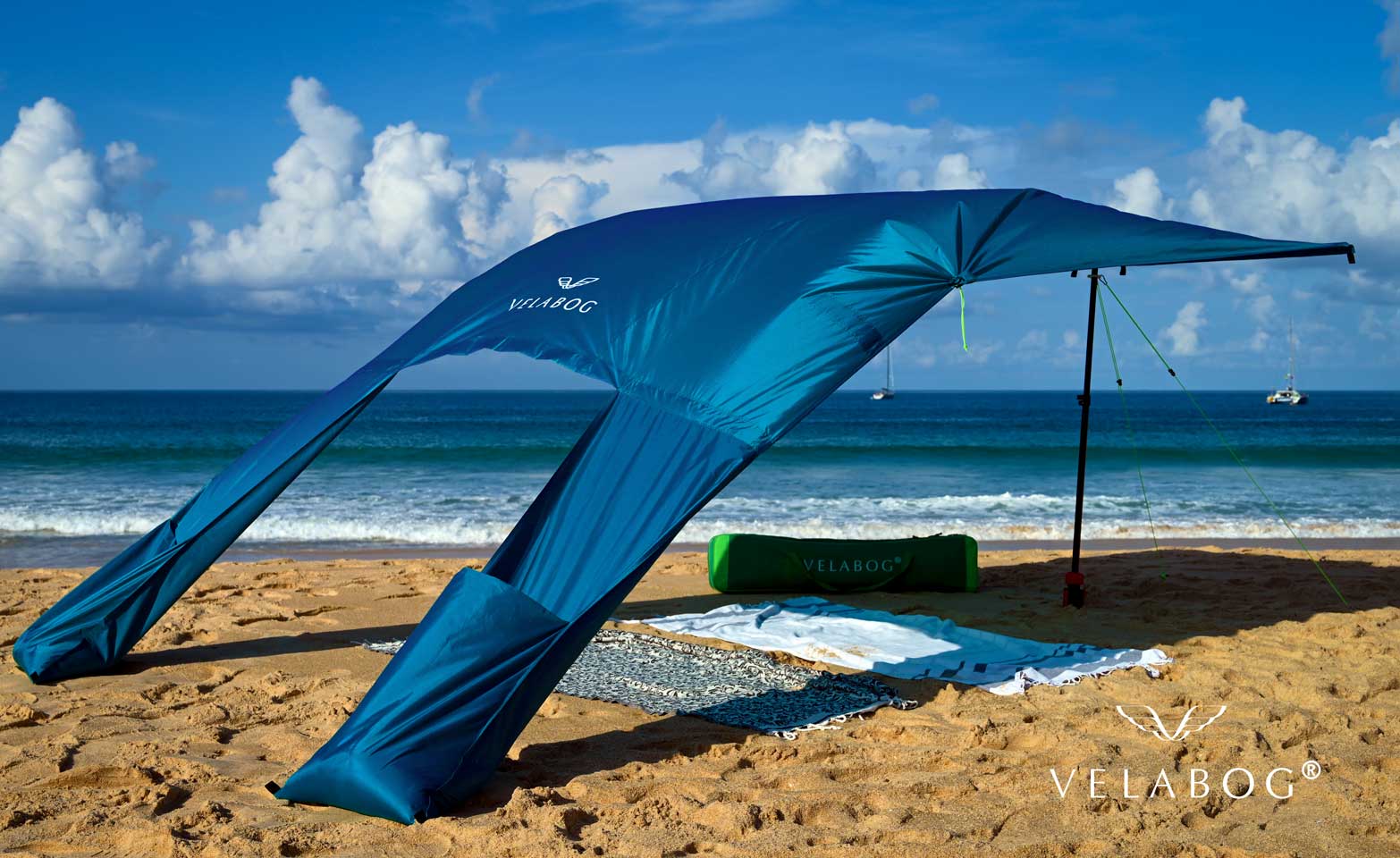 Toldo playa Velabog Breeze azul. La mejor refugio y protección solar en la playa. Mucha más sombra en comparación con sombrillas de playa. Mucho más aire en comparación con carpas de playa. Opción de montaje desde viento ligero a fuerte. Vista inclinada desde atrás.
