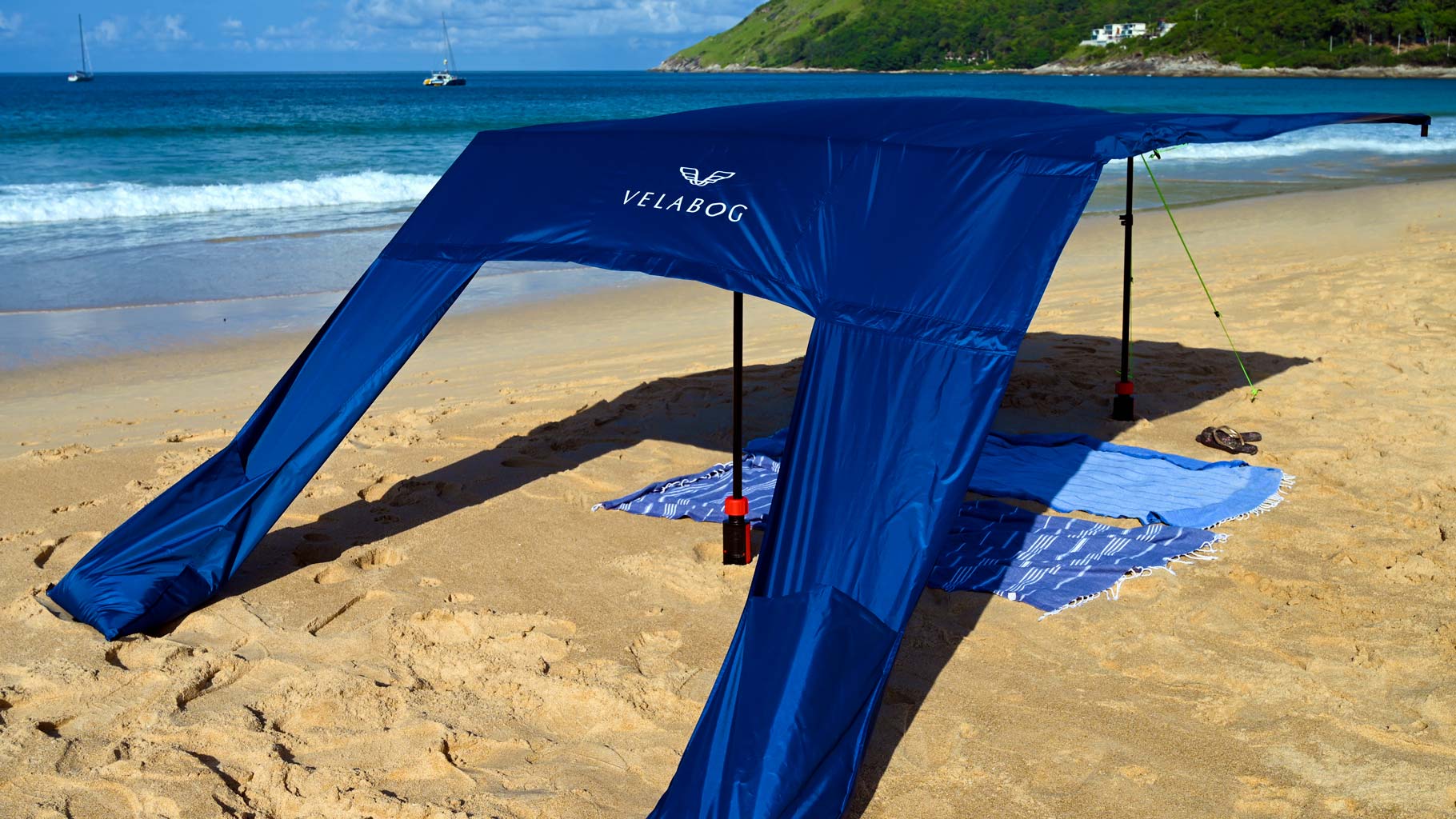 Voile d’ombrage tente plage Velabog Breeze bleu nuit avec kit d’extension sur une plage balayée par des rafales de vent. Voile d’ombrage, parasol et tente de plage en un seul produit. Offre beaucoup d’ombre par tout type de vent, qu’il soit faible, fort ou en rafales. Vue de dos.